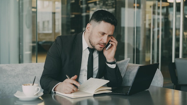 Hombre de negocios caucásico barbudo que usa una computadora portátil hablando por teléfono inteligente y escribiendo información comercial en el café durante el almuerzo