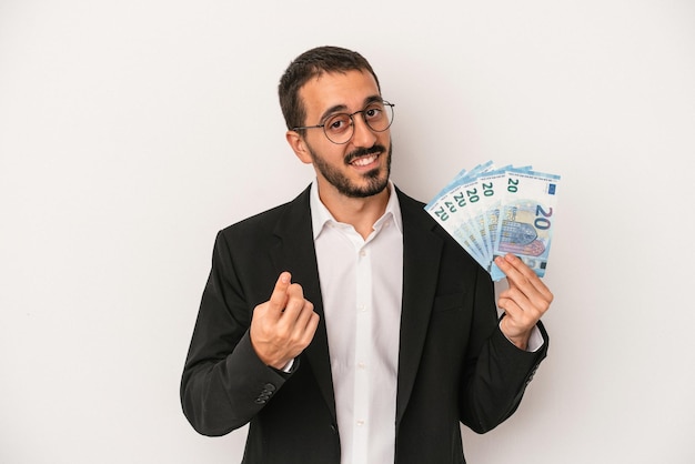 Hombre de negocios caucásico joven con billetes aislados sobre fondo blanco apuntando con el dedo a usted como si invitando a acercarse.