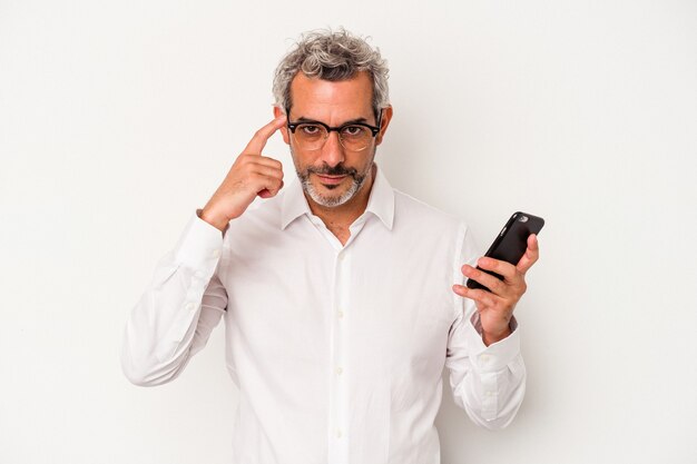 Hombre de negocios caucásico de mediana edad sosteniendo un teléfono móvil aislado sobre fondo blanco señalando la sien con el dedo, pensando, centrado en una tarea.