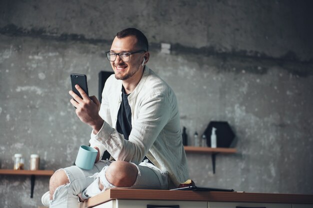 Hombre de negocios caucásico en ropa de mezclilla blanca está usando un teléfono y auriculares mientras toma un café en casa