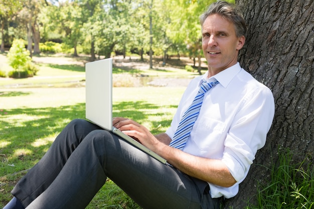 Hombre de negocios con la computadora portátil apoyándose en el árbol