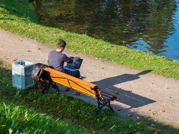 Un hombre de negocios con una computadora portátil trabaja sentado en un banco en un parque de otoño.