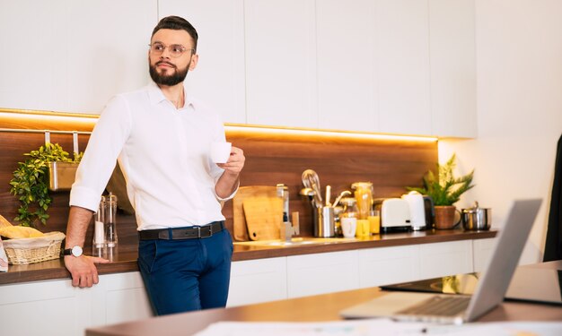 Hombre de negocios confiado barbudo relajado en ropa casual elegante bebidas huele café y reflexivo en la cocina doméstica