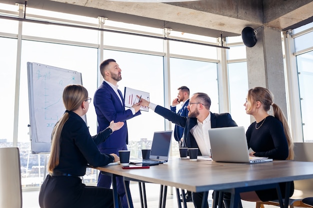 Hombre de negocios confiado hace una presentación de un nuevo proyecto en la sala de juntas en una reunión de la empresa. Hermosos auditores hablan con diferentes socios sobre el negocio usando una pizarra y gráficos.