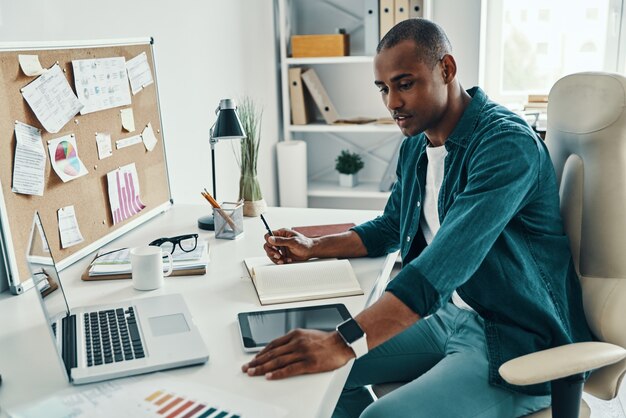 Hombre de negocios confiado. Hombre africano joven pensativo en camisa usando la computadora portátil mientras está sentado en la oficina