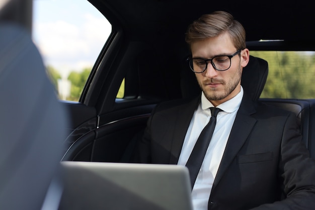 Hombre de negocios confiado pensativo que se sienta en el coche de lujo y que usa su computadora portátil.