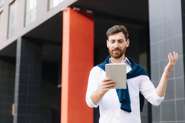 Hombre de negocios confiado en ropa de moda con videollamada en tableta digital mientras está de pie cerca del edificio de oficinas moderno. Concepto de conversación y tecnología.