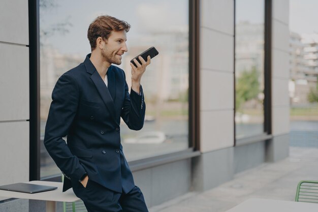 Hombre de negocios confiado con smartphone usando la aplicación de asistente de voz mientras se apoya en la mesa de café al aire libre