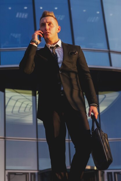 Hombre de negocios contemplativo joven con maletín deja edificio comercial cerca de la empresa corporativa y llamando al teléfono móvil, estilo de vida. Banquero masculino saliendo de la oficina ubicada en el distrito financiero