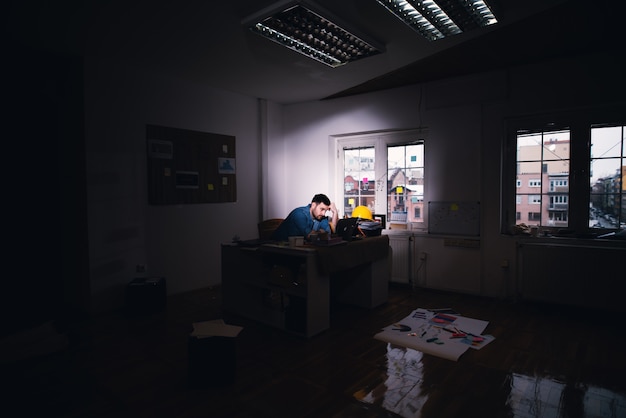 Hombre de negocios decepcionado cansado joven que busca la solución del problema en una computadora portátil mientras se queda después del horario de trabajo regular en la oficina oscura.