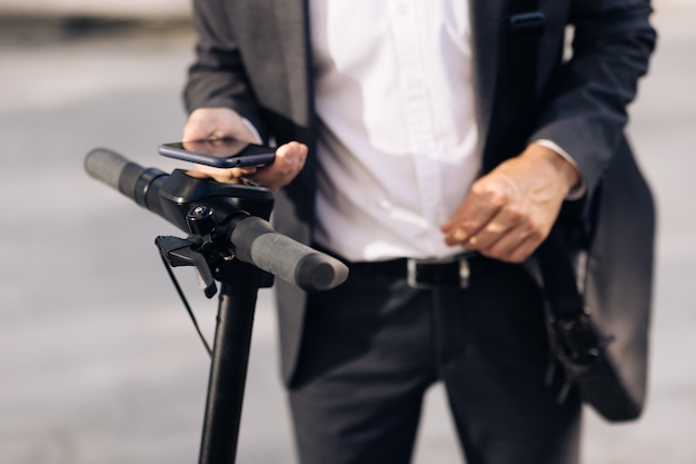 Hombre de negocios desconocido con traje clásico se acerca a un scooter eléctrico y usa la aplicación de teléfono móvil