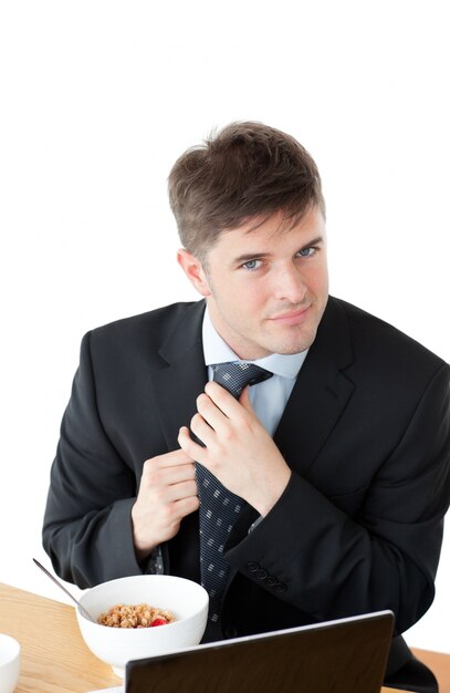 Hombre de negocios elegante desayunando y tocando su corbata frente a su computadora portátil