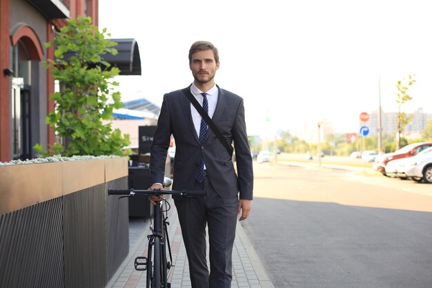 Hombre de negocios elegante joven vestido con traje caminando con una bicicleta en una calle de la ciudad.