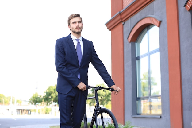 Hombre de negocios elegante joven vestido con traje caminando con una bicicleta en una calle de la ciudad.