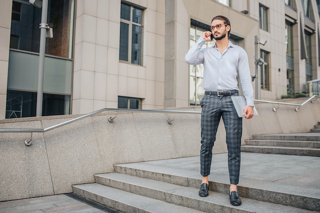 Hombre de negocios elegante serio y concentrado se encuentra y plantea. Él sostiene la computadora portátil en la mano y el teléfono cerca de la oreja. Él mira hacia adelante.