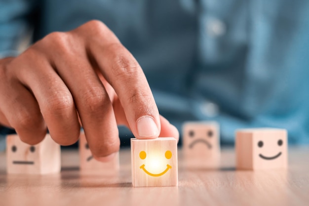 El hombre de negocios elige un icono de emoticono de sonrisa que se enfrenta a un símbolo feliz en un bloque de madera, servicios y concepto de encuesta de satisfacción del cliente