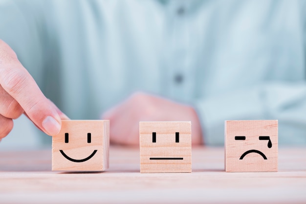 El hombre de negocios elige un icono de emoticono de sonrisa que se enfrenta a un símbolo feliz en un bloque de madera, servicios y concepto de encuesta de satisfacción del cliente