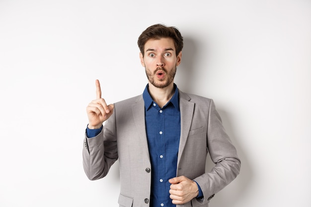 Hombre de negocios emocionado en traje lanzando una buena idea, levantando el dedo en el signo de eureka y mirando divertido, diciendo sugerencia, de pie sobre fondo blanco.