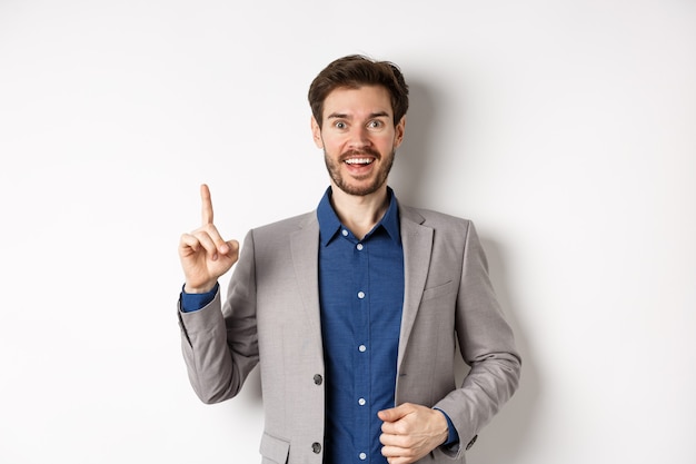 Hombre de negocios emocionado en traje lanzando una buena idea, levantando el dedo en el signo de eureka y mirando divertido, diciendo sugerencia, de pie sobre fondo blanco.