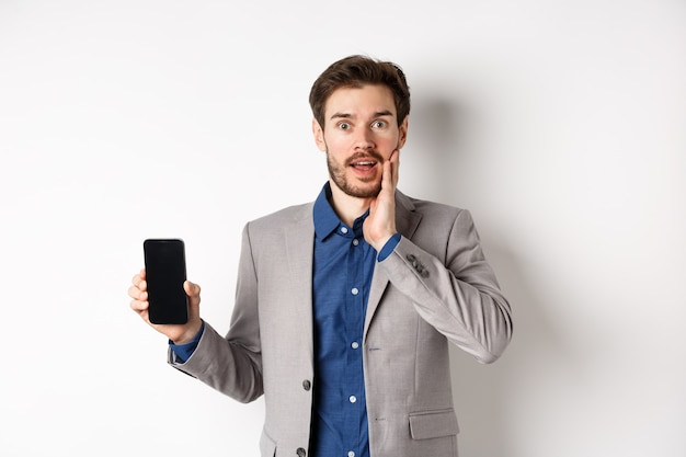 Hombre de negocios emocionado en traje mostrando la pantalla vacía del teléfono inteligente y jadeando asombrado, demostrar la aplicación, de pie sobre fondo blanco.