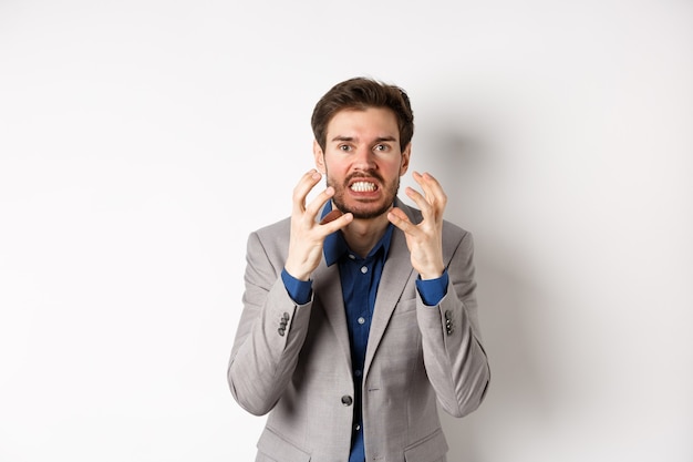 Hombre de negocios enojado y molesto quiere matar a alguien, apretando las manos y los dientes, mirando enojado a la cámara, yendo a estrangular a la persona, de pie sobre un fondo blanco.