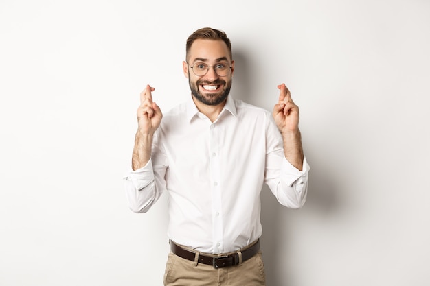 Hombre de negocios esperanzado sonriendo y cruzando los dedos para la buena suerte, pidiendo un deseo, de pie sobre fondo blanco.