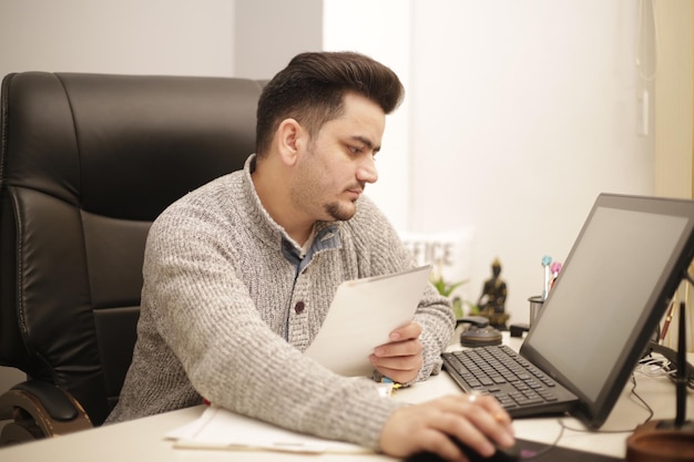 Un hombre de negocios está trabajando en una computadora mientras sostiene el papel en la mano.