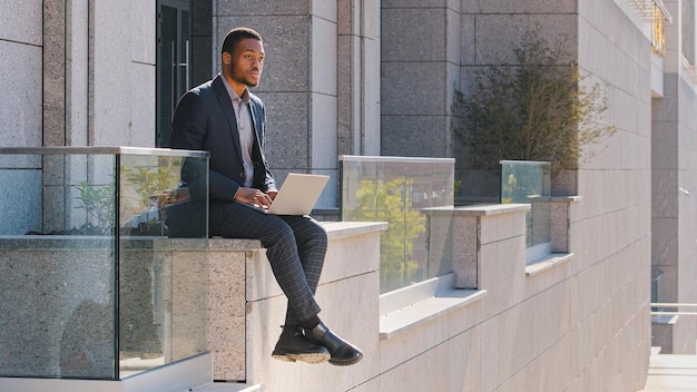 Hombre de negocios étnico reflexivo Gerente de hombre afroamericano independiente sentado al aire libre escribiendo en una computadora portátil preocupado por el tipo de trabajador problemático pensando en una decisión difícil mirando hacia otro lado la estrategia de planificación