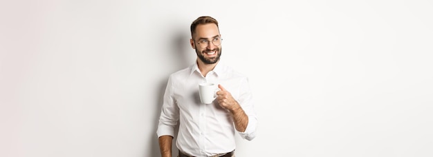 Hombre de negocios exitoso bebiendo café mirando hacia los lados con una sonrisa satisfecha de pie sobre ba blanca