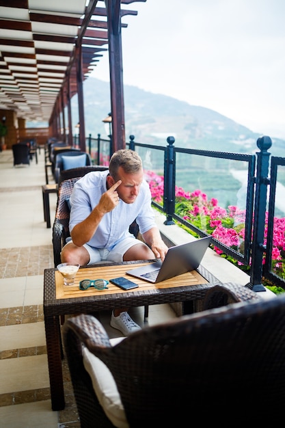 Un hombre de negocios exitoso está sentado en una mesa con una computadora portátil, está trabajando de vacaciones. Vacaciones y trabajo a distancia. El chico está sentado en un restaurante en una terraza con vista panorámica.