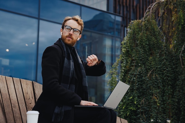 Hombre de negocios exitoso en el fondo del centro de negocios Joven pelirrojo con un elegante abrigo Auriculares inalámbricos con una computadora portátil