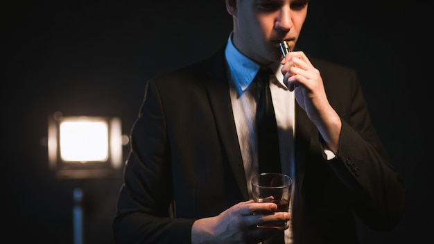 Hombre de negocios exitoso fumando cigarrillo electrónico y tomando una copa. Relajarse y disfrutar de la vida después del trabajo duro.