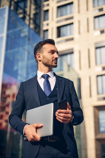 Hombre de negocios exitoso pasar tiempo fuera del gran centro de negocios con una computadora portátil y un teléfono móvil en las manos