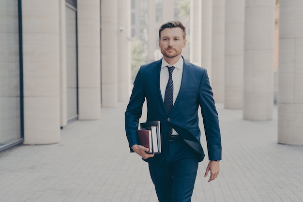 Hombre de negocios exitoso seguro en ropa formal con computadora portátil, cuaderno y libro en mano caminando por las calles de la ciudad con fondo urbano, apresurándose en la reunión de negocios con socio o cliente