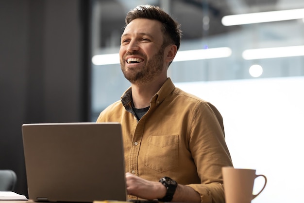 Un hombre de negocios exitoso usando una computadora portátil y riendo mirando a un lado en el lugar de trabajo