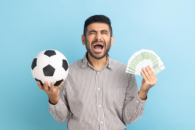 Hombre de negocios extremadamente feliz sosteniendo un balón de fútbol y fanático de los billetes en euros apuestas deportivas gran victoria regocijándose gritando con camisa a rayas Estudio interior aislado en fondo azul