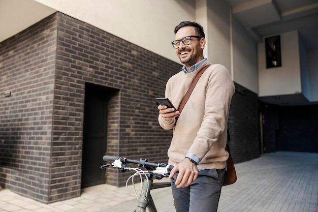 Un hombre de negocios feliz e inteligente está empujando su bicicleta en la calle y usando el teléfono en la calle