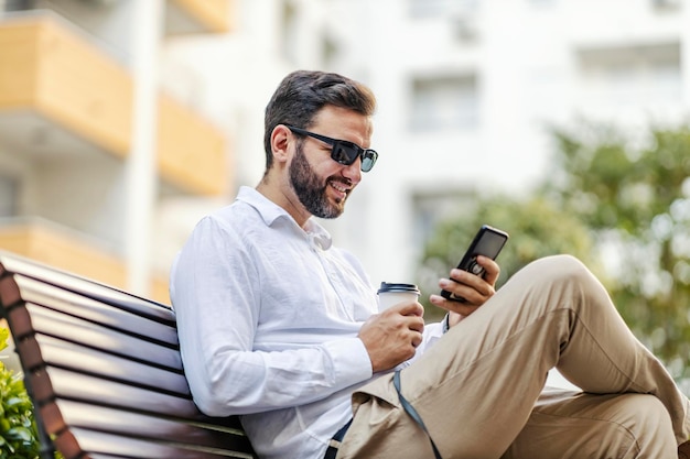 Un hombre de negocios feliz con gafas de sol está sentado en un banco en el parque con café y usando aplicaciones