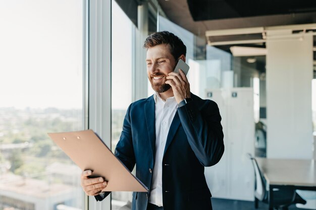 Hombre de negocios feliz hablando por teléfono inteligente y revisando papeles de pie en el espacio libre interior de la oficina moderna