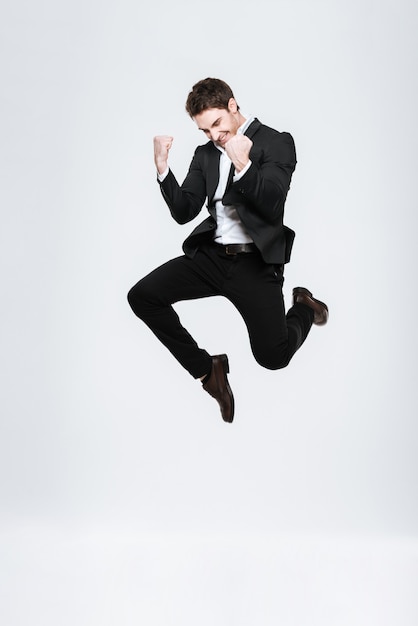Foto hombre de negocios feliz de longitud completa en traje negro saltando y celebrando el éxito aislado en una pared blanca