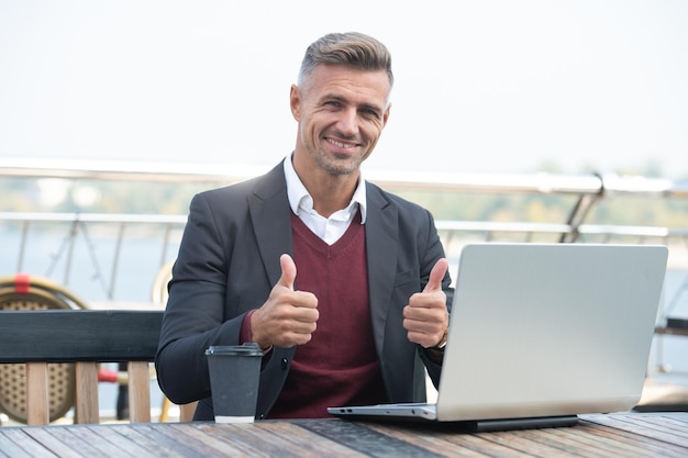 Hombre de negocios feliz muestra los pulgares haciendo gesto de aprobación trabajando en la computadora portátil afuera, pulgares hacia arriba.