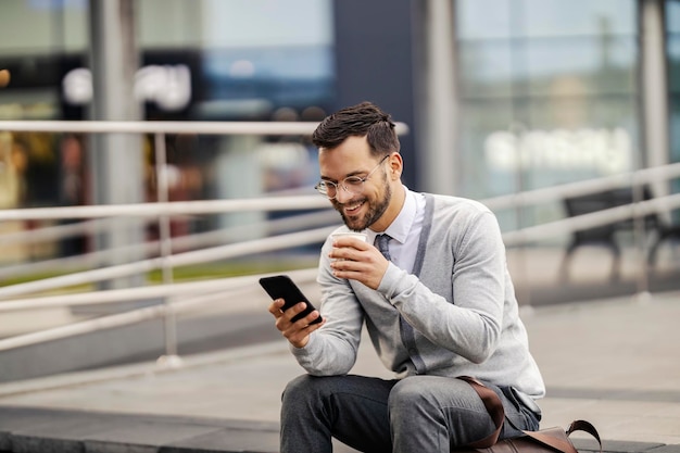 Un hombre de negocios feliz se sienta afuera bebe café y sonríe al teléfono en su descanso del trabajo