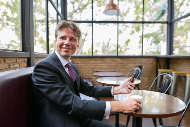 Hombre de negocios feliz sonriendo mientras sostiene el teléfono móvil y el café