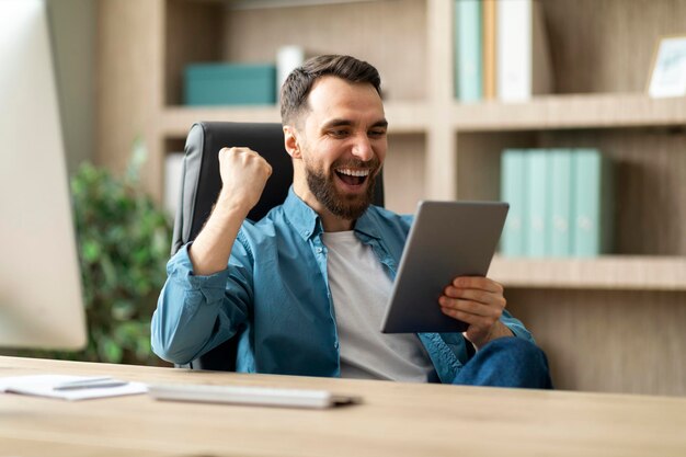 Hombre de negocios feliz con tableta digital celebrando el éxito en la oficina sentado en el lugar de trabajo