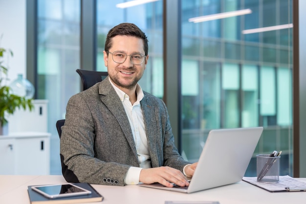 Un hombre de negocios feliz trabajando en una computadora portátil en la oficina mirando a la cámara con una sonrisa