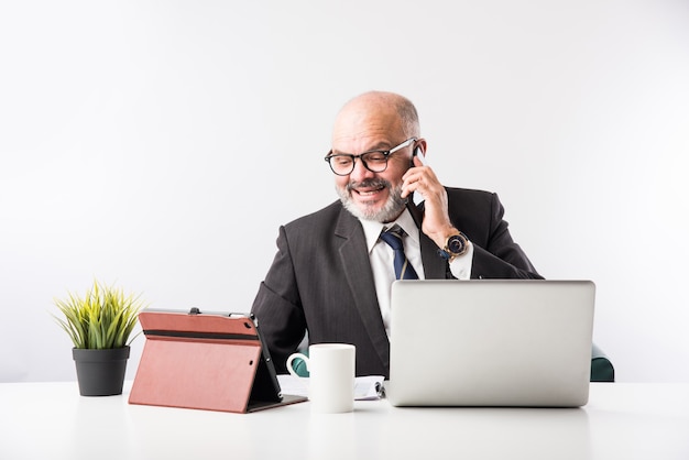 Hombre de negocios financiero senior indio asiático sentado en su estación de trabajo o escritorio frente a una computadora, computadora portátil y tableta. Hablar por teléfono mientras hace algunos trámites