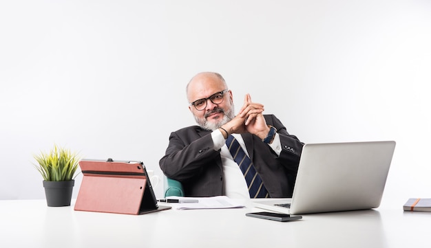 Hombre de negocios financiero senior indio asiático sentado en su estación de trabajo o escritorio frente a una computadora, computadora portátil y tableta. Hablar por teléfono mientras hace algunos trámites