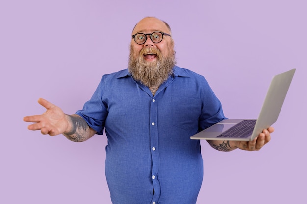 Hombre de negocios gordo alegre en camisa ajustada con gafas sostiene portátil sobre fondo púrpura