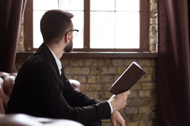 Hombre de negocios guapo y confiado con traje inteligente y anteojos leyendo un libro o revisando los informes sobre el trabajo de sentarse en el sofá de la oficina