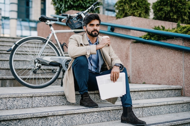 Hombre de negocios guapo joven sentado en las escaleras en las calles de la ciudad con bicicleta, mientras bebe café para ir y leer el periódico. Protección del medio ambiente y concepto de transporte ecológico.